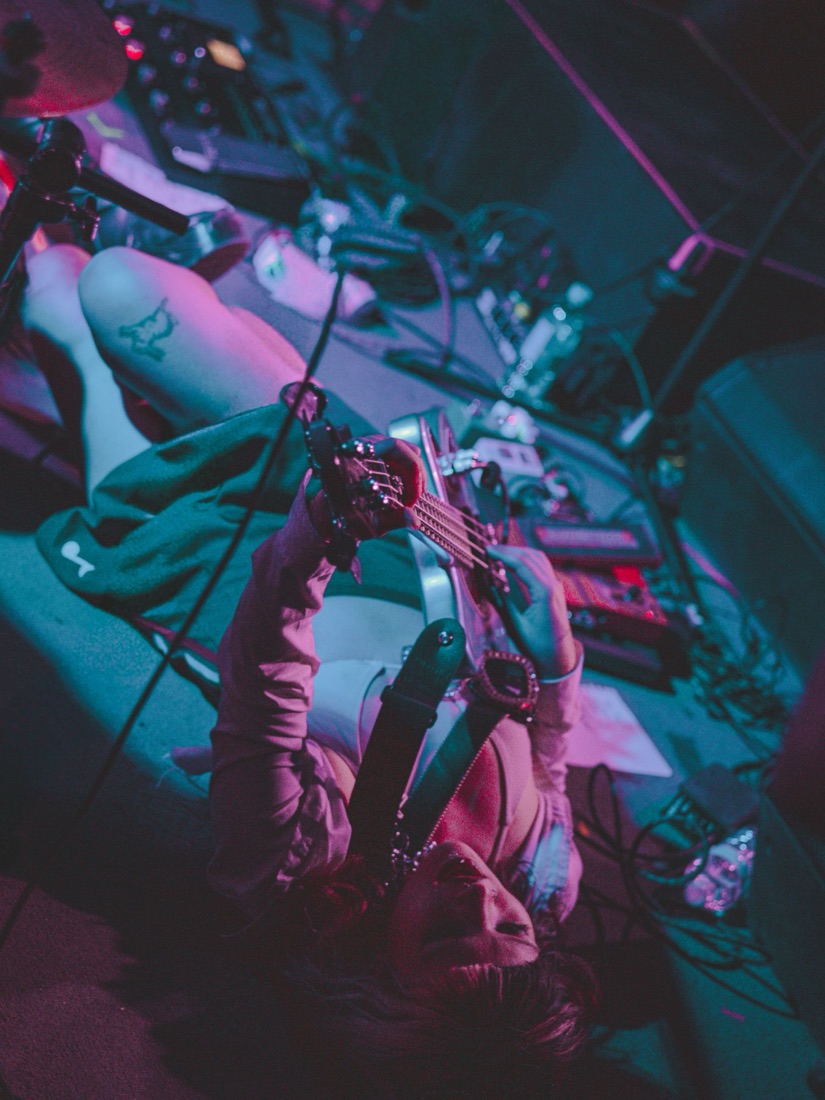 Bassist lying on the stage floor among pedals, shot from above in pink and teal