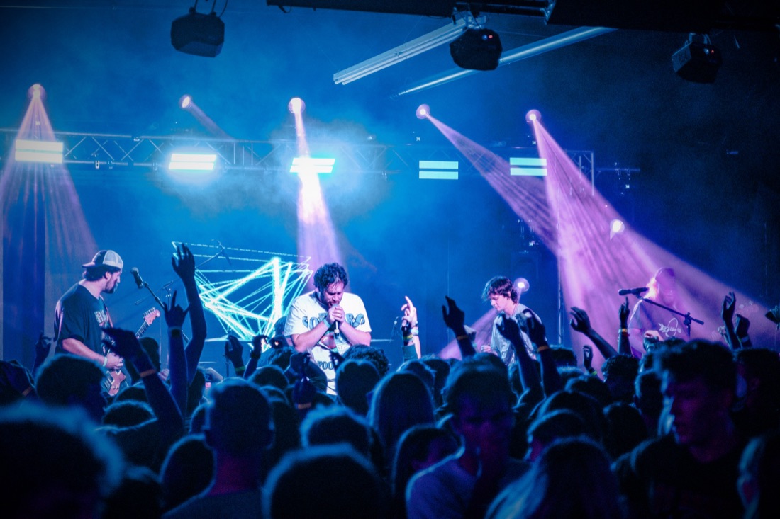 Full band on stage under blue and purple beams with hands raised in the crowd